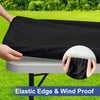A hand is holding a black elastic edge and wind proof table cover over a white folding table outdoors.