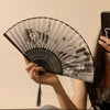 A black and white antique style bamboo folding hand fan is held by a person against a light background.