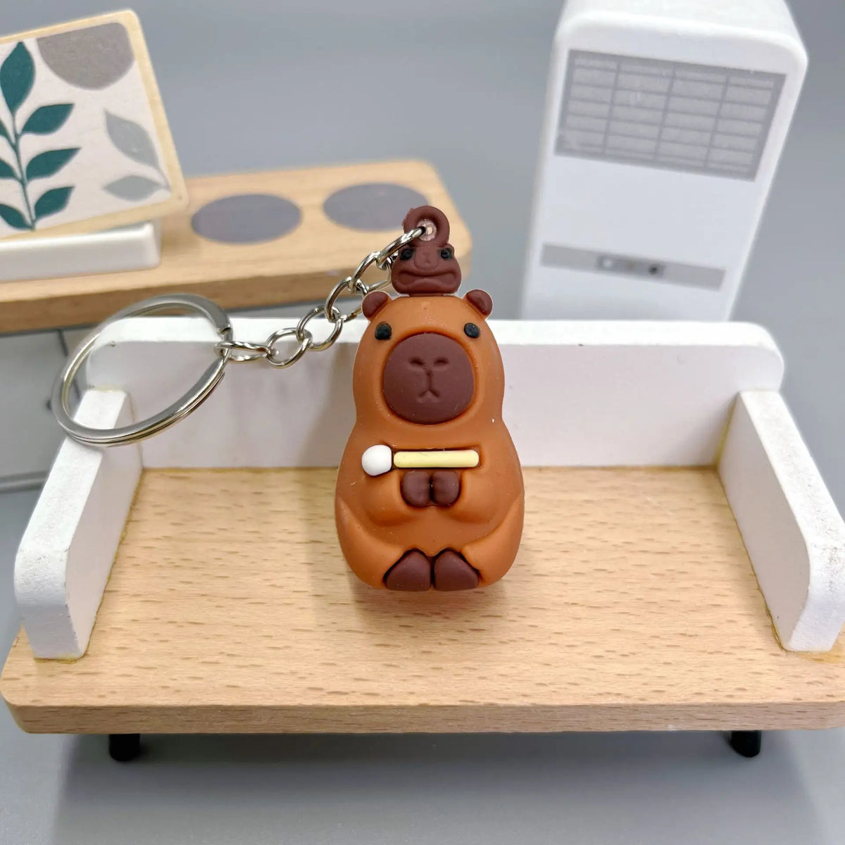 Pendentif de capybara orange en forme de chapeau, suspendu à une chaîne argentée, exposé sur une étagère en bois.