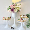 A white pedestal vase holds a lush arrangement of pink, purple, and cream roses and greenery for a centerpiece display.