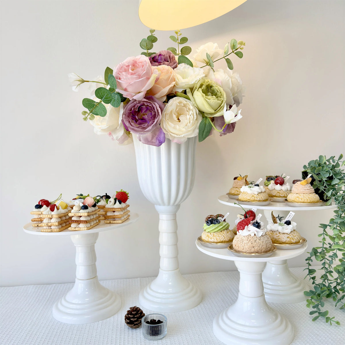 A white pedestal vase holds a lush arrangement of pink, purple, and cream roses and greenery for a centerpiece display.