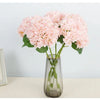 A bouquet of pink artificial hydrangea flowers in a clear glass vase on a light surface.