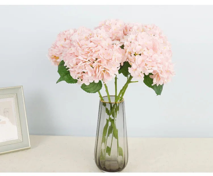 A bouquet of pink artificial hydrangea flowers in a clear glass vase on a light surface.