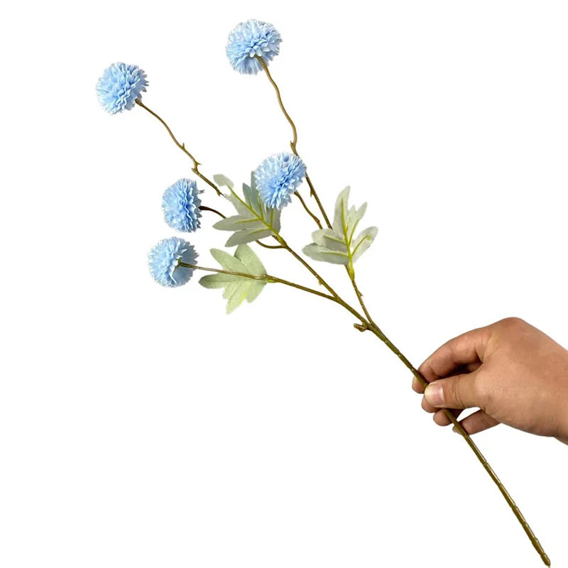Una mano sostiene una ramita de flores artificiales de diente de león azules con hojas verdes.