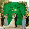 A wedding photo featuring a couple standing in front of a green fabric backdrop decorated with white flowers and the word Lov