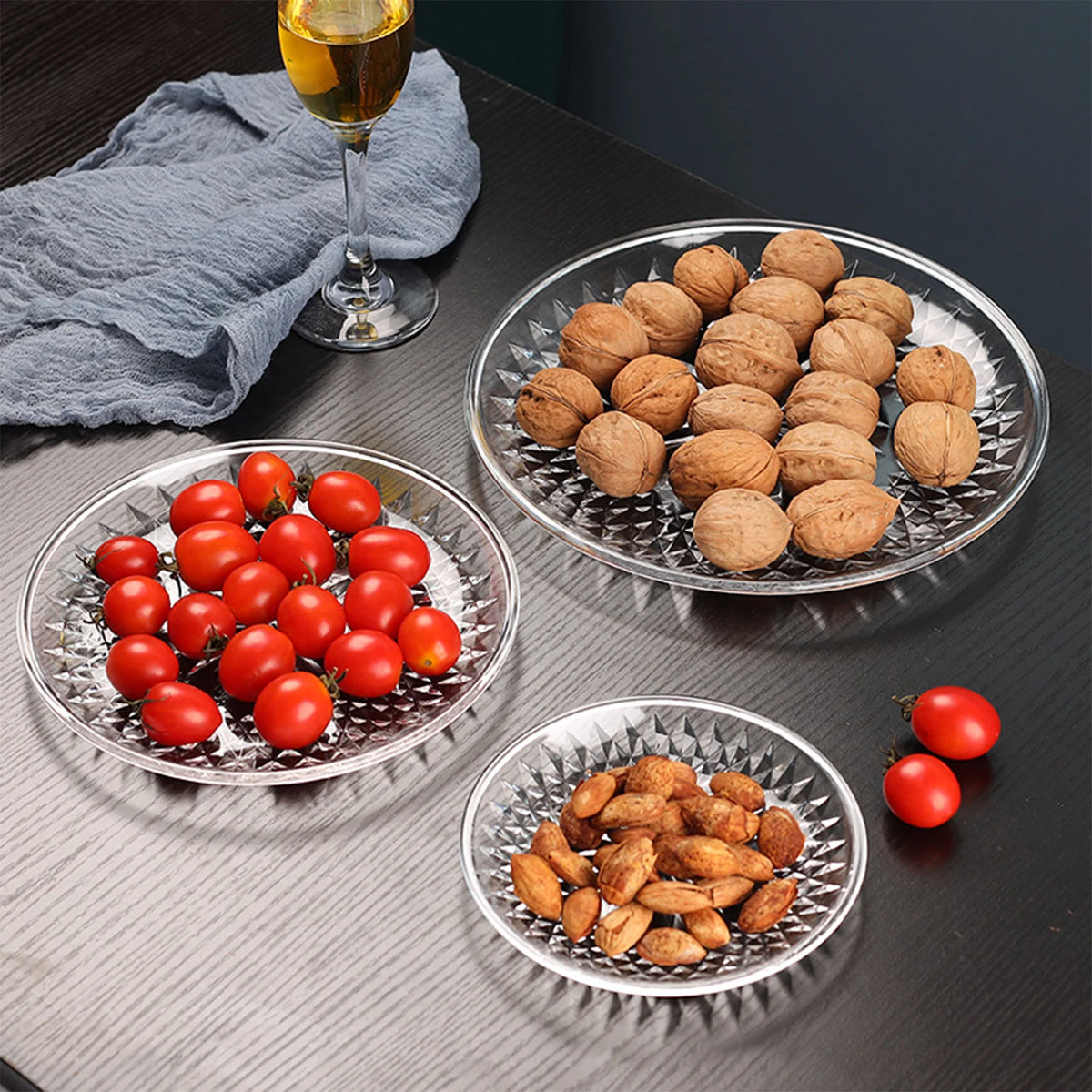 Clear acrylic serving plates filled with various nuts and cherry tomatoes on a dark surface.