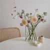 A clear acrylic book vase filled with dried flowers and greenery on a white table.