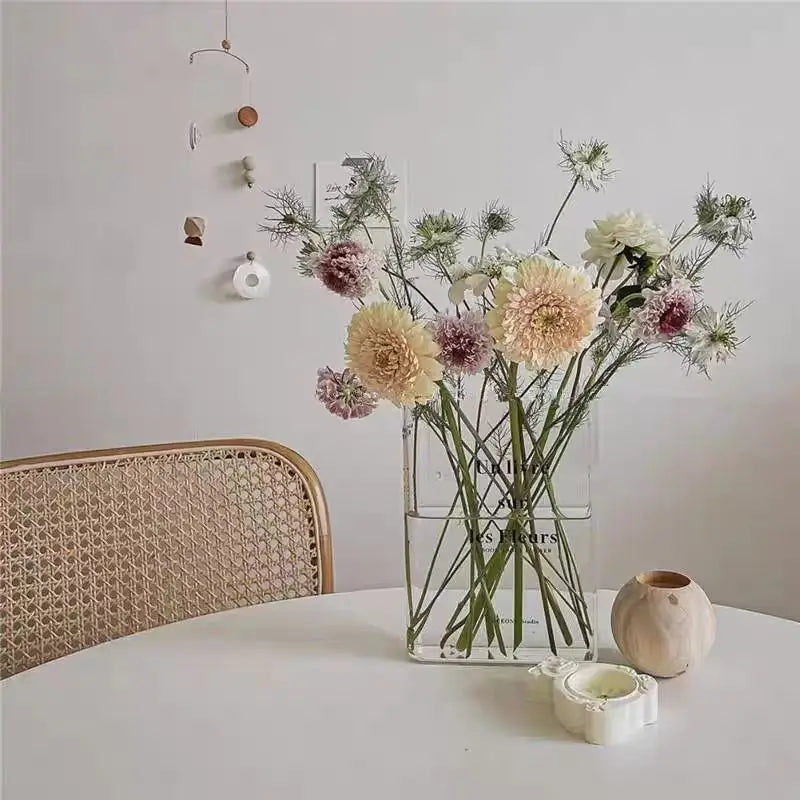 A clear acrylic book vase filled with dried flowers and greenery on a white table.
