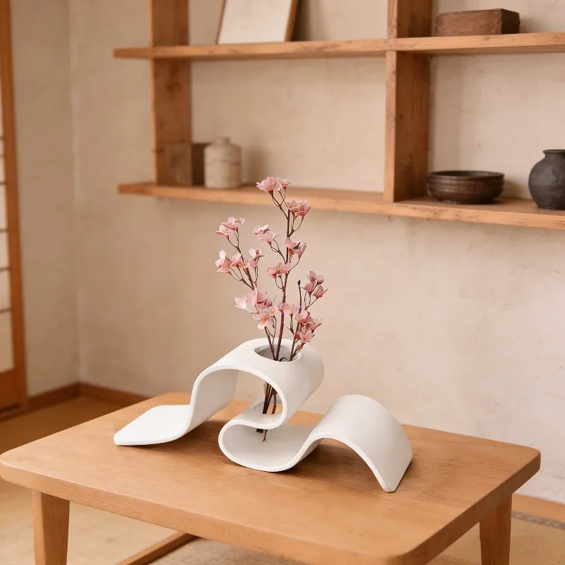 Jarrón curvo blanco moderno con ramas rosadas sobre una mesa de madera en un interior luminoso.
