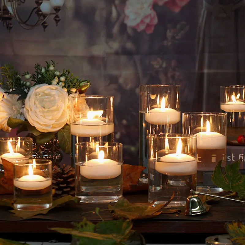 A collection of white candles in clear glass holders arranged on a dark surface with greenery and natural elements.