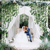 A wedding or event setup featuring a white archway decorated with lush green ivy vines and sheer white fabric.