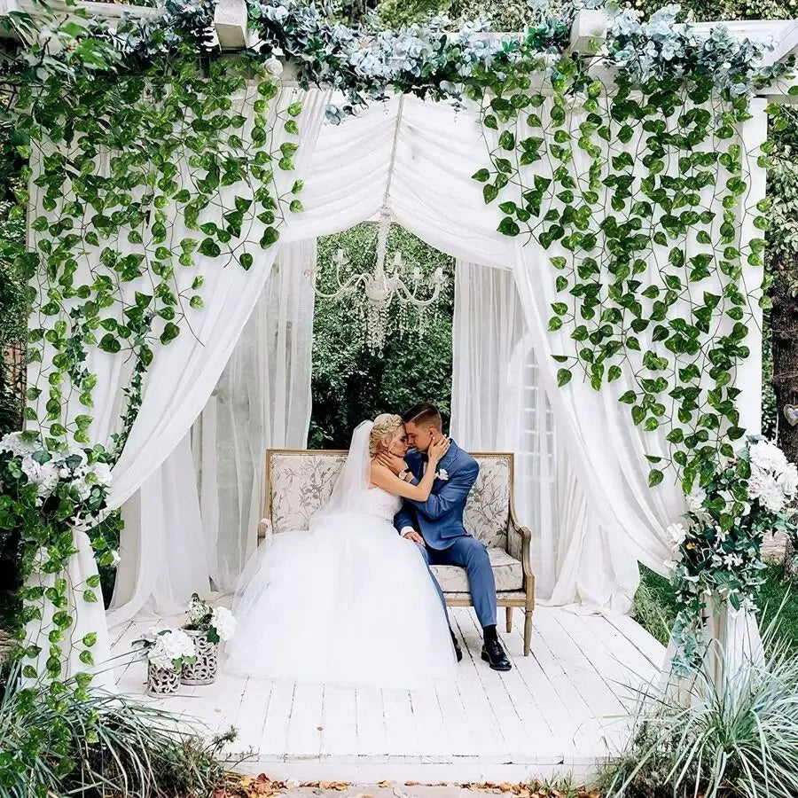 A wedding or event setup featuring a white archway decorated with lush green ivy vines and sheer white fabric.