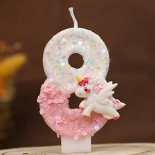 Vela blanca con forma de 8, decorada con purpurina rosa y una pequeña figura de unicornio blanco.
