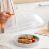 A hand holds a transparent microwave splatter guard with an orange handle over a plate of food.