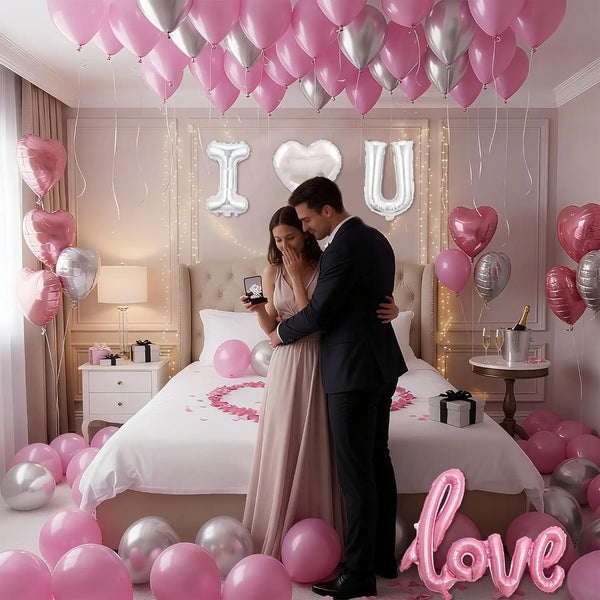 A couple stands in a bedroom surrounded by pink and silver heart foil balloons on a bed.