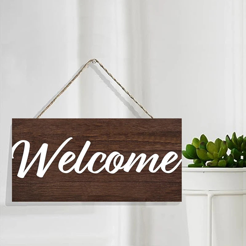 A rustic wooden welcome sign with white script text on a dark brown background, hanging on a gold wire.