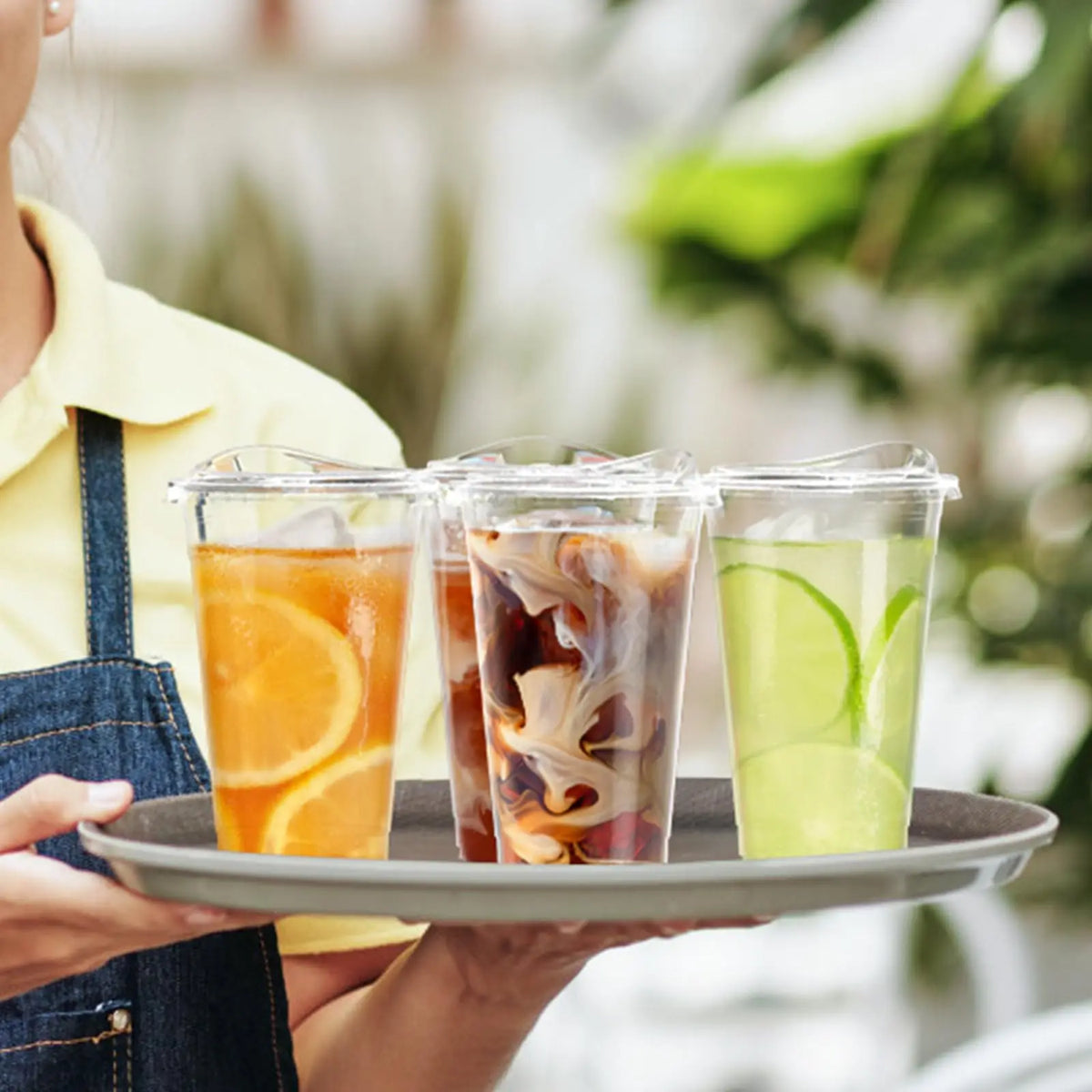A hand holds a tray of three clear plastic cups filled with iced beverages, including orange juice and green lemonade.