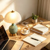 A wooden desk features a ceramic table lamp, a laptop, and notebooks under bright natural light.