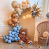 A decorative arch backdrop made of light beige balloons and a wooden bear figure for a party or event.