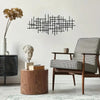 A black geometric metal wall art sculpture hangs on a white wall above a wooden table and an upholstered chair.