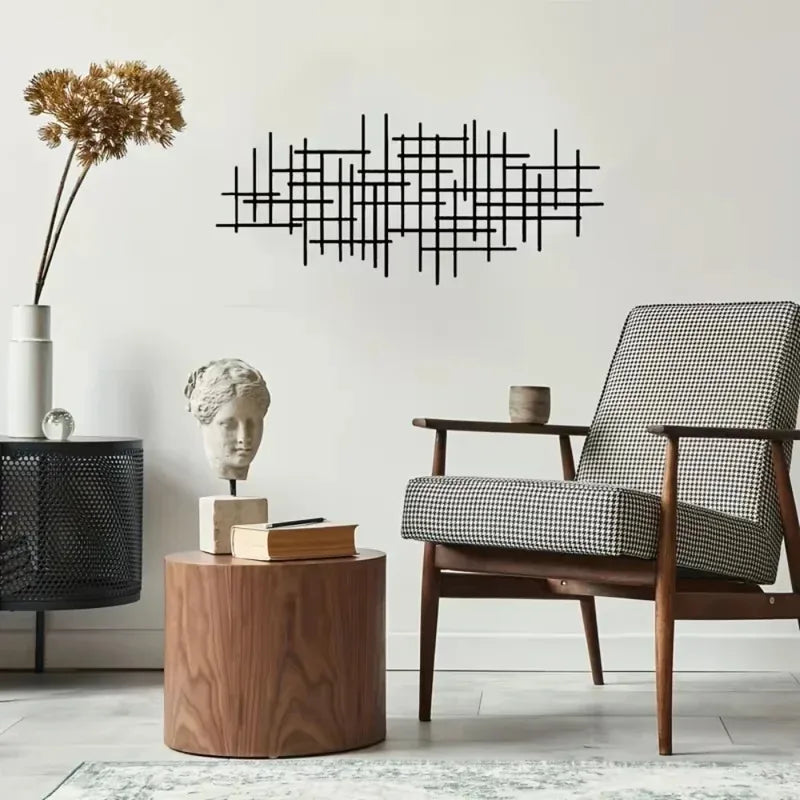 A black geometric metal wall art sculpture hangs on a white wall above a wooden table and an upholstered chair.