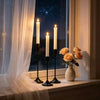 A set of three tall white taper candles in black holders displayed on a windowsill with a window view.