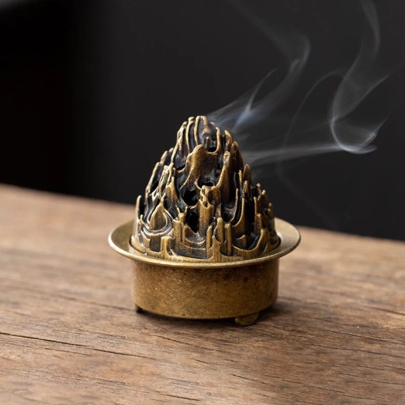 A vintage brass incense burner holder with a textured top is emitting smoke on a wooden surface.