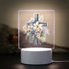 A 3D LED night light featuring a wooden cross adorned with white and purple flowers on a square base.