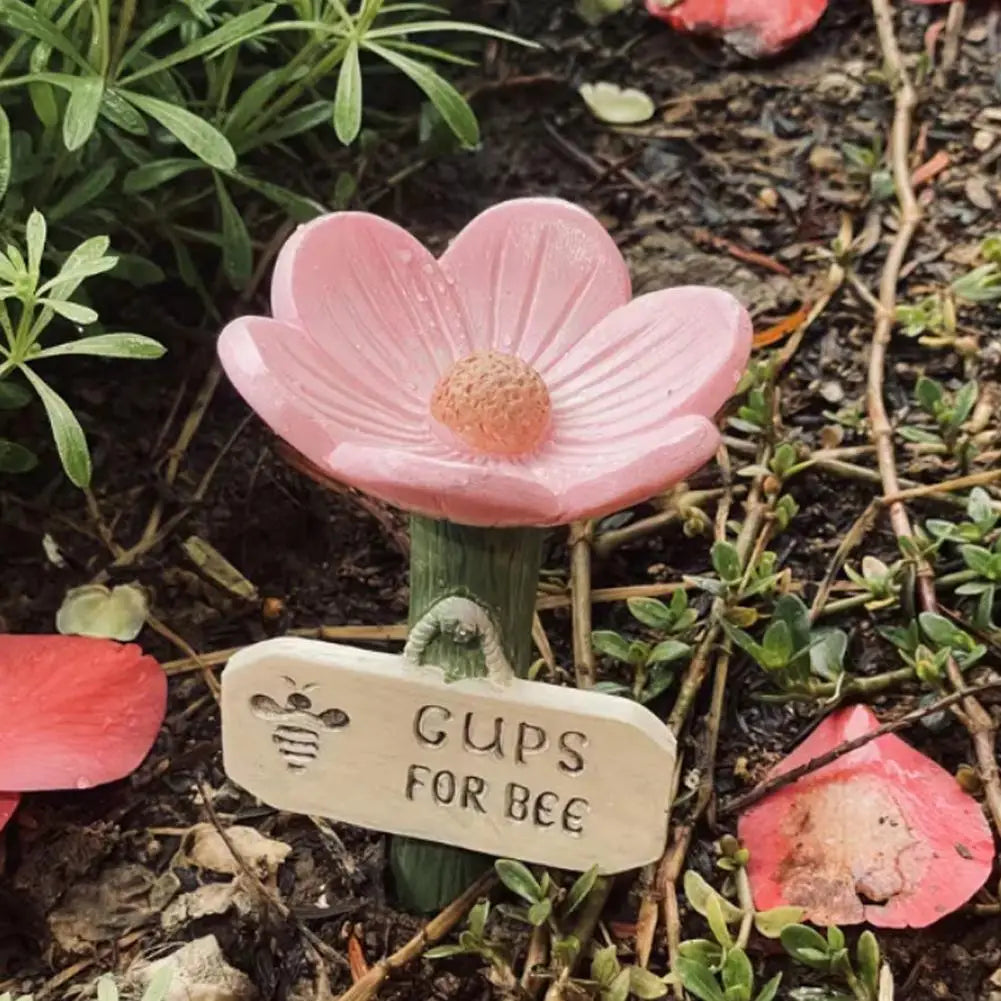 A pink flower with a bee-shaped tag is shown growing in dark soil.