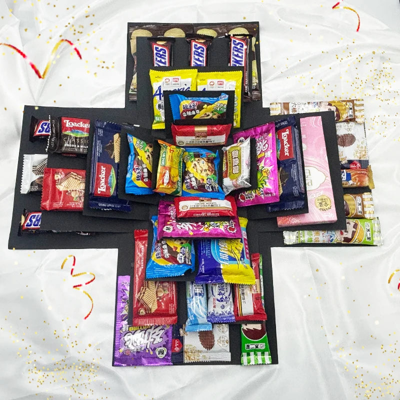 A black gift box is filled with various colorful party snacks and candies arranged in a cross shape.