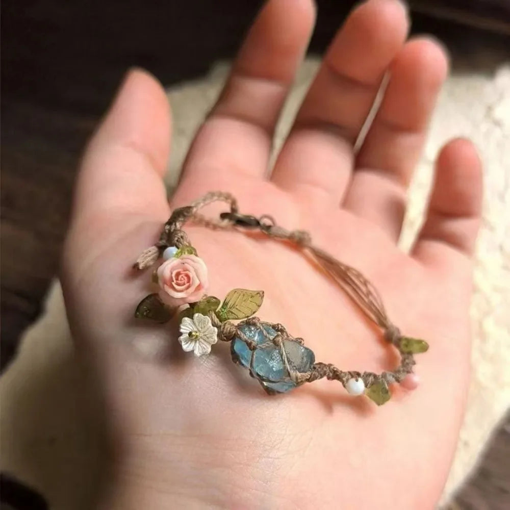 A hand holds a raw aquamarine macrame bracelet featuring a rose accent and small blue stones.