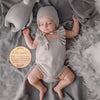A baby wearing a knitted outfit is lying on a gray blanket next to a wooden name announcement sign.