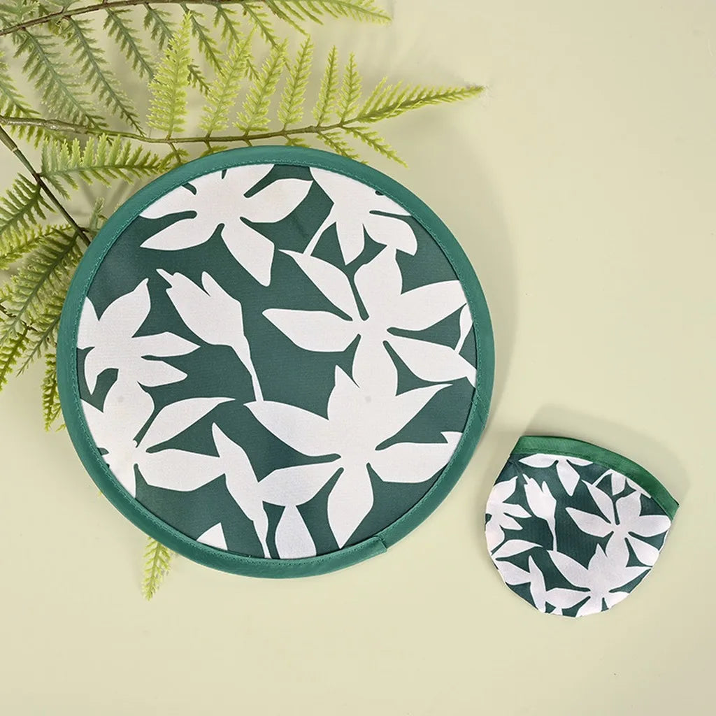 A round, dark green fabric hand fan with a white floral pattern is shown against a light background.