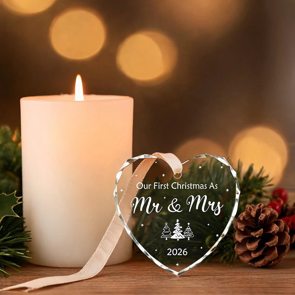 A glowing candle next to a heart-shaped crystal ornament engraved with Our First Christmas As Mr & Mrs 2026 surrounded by pin