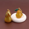 A gold-colored pear-shaped fruit keychain charm with a green leaf detail resting on a white circular surface.