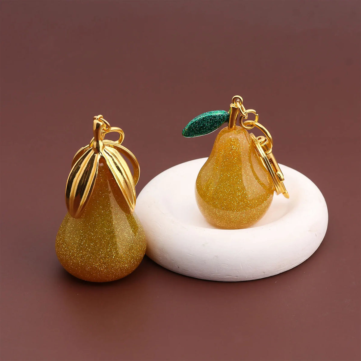 A gold-colored pear-shaped fruit keychain charm with a green leaf detail resting on a white circular surface.