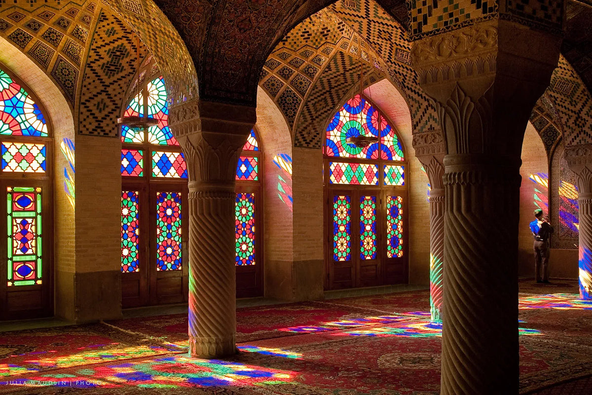 Interior view of an Islamic mosque with arched windows and columns, featuring colorful stained glass and patterned rugs.