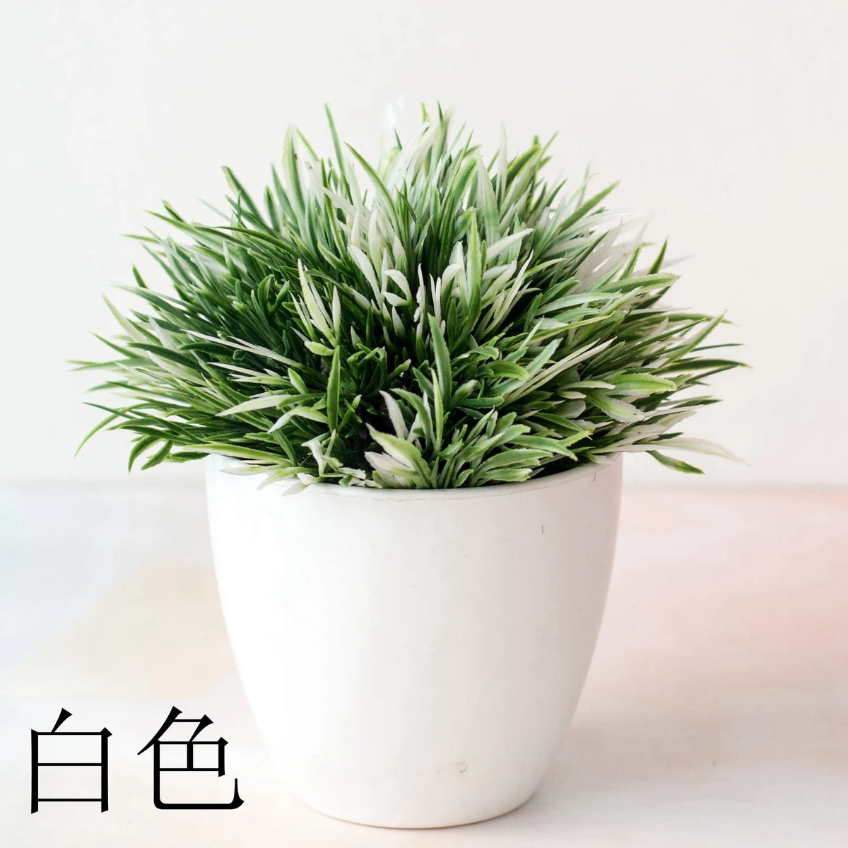 Un pot de bonsaï artificiel avec du feuillage vert clair et blanc repose sur une surface blanche.
