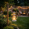 A decorative metal candlestick lantern hangs outdoors in a garden setting surrounded by greenery and string lights.