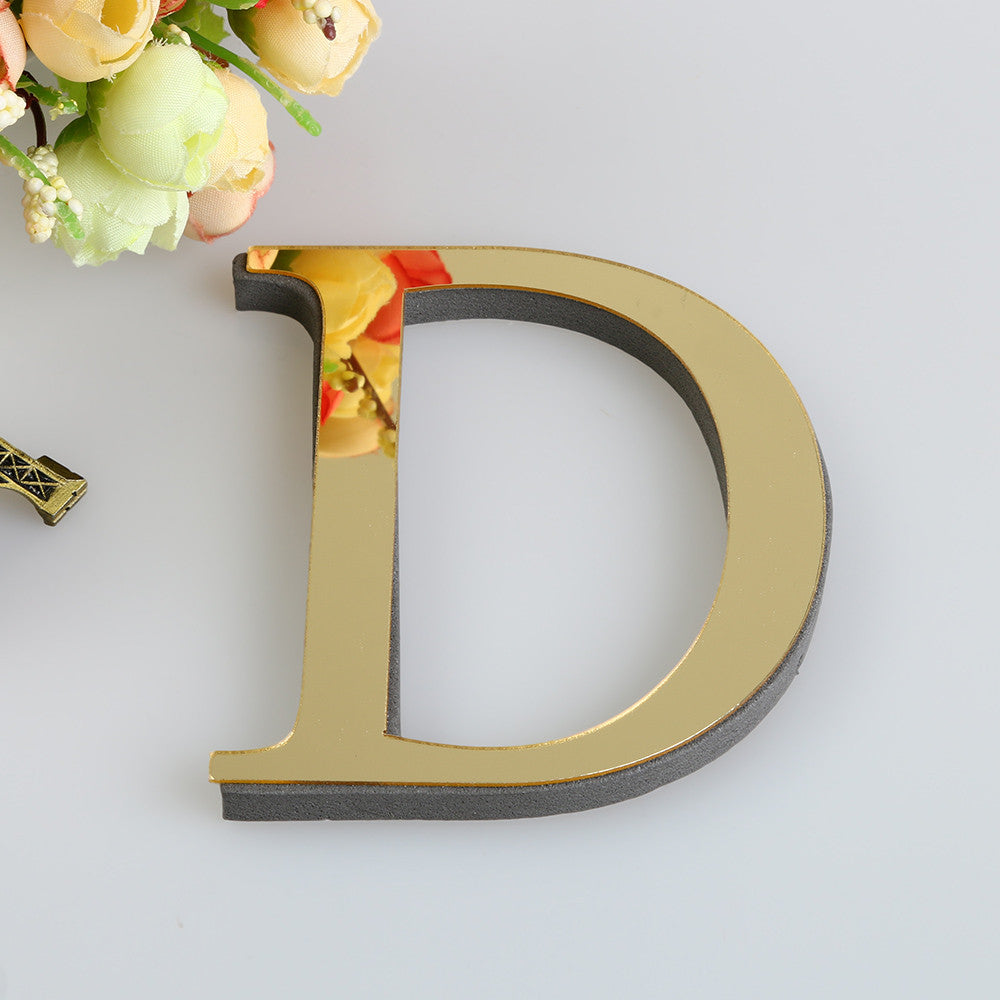 A gold letter D cutout for a mirror wall decal set, placed next to a bouquet of light yellow and green flowers.
