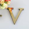 A gold letter V cutout with a mirrored finish is placed next to a bouquet of light yellow and pink flowers.