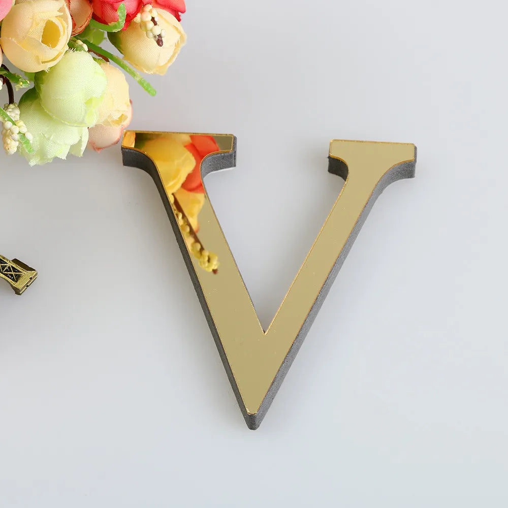 A gold letter V cutout with a mirrored finish is placed next to a bouquet of light yellow and pink flowers.