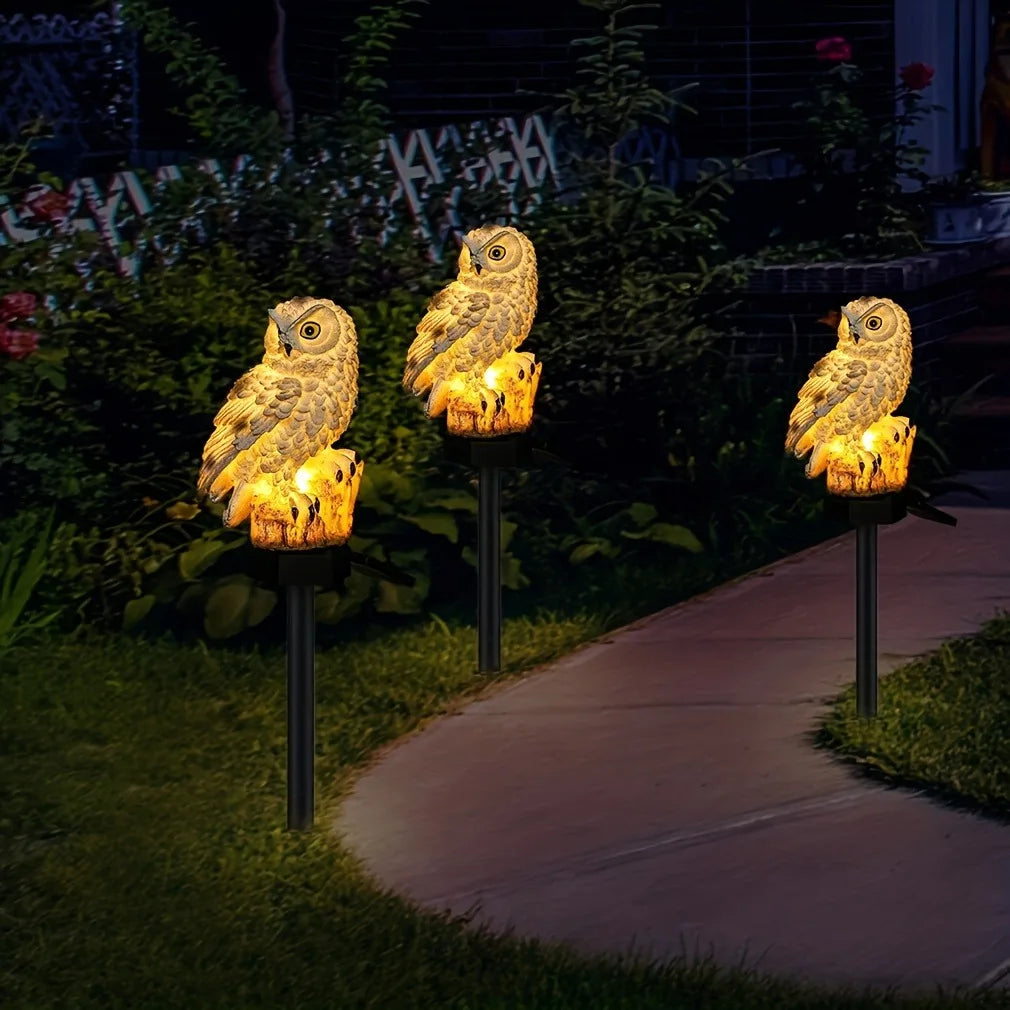 A solar-powered owl garden stake light with an owl figure illuminates a pathway in a garden setting.