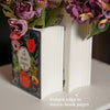 A white ceramic book-shaped vase decorated with floral elements and a ridged edge.