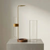 Un haut brûleur d'encens en laiton élancé avec un cylindre de verre et un verre à eau transparent sur une surface gris clair.