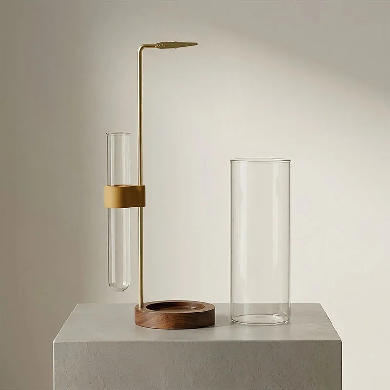 Un haut brûleur d'encens en laiton élancé avec un cylindre de verre et un verre à eau transparent sur une surface gris clair.