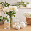 A clear glass vase holds white peonies next to a wooden shelf with ceramic teapot and small white decorative items.