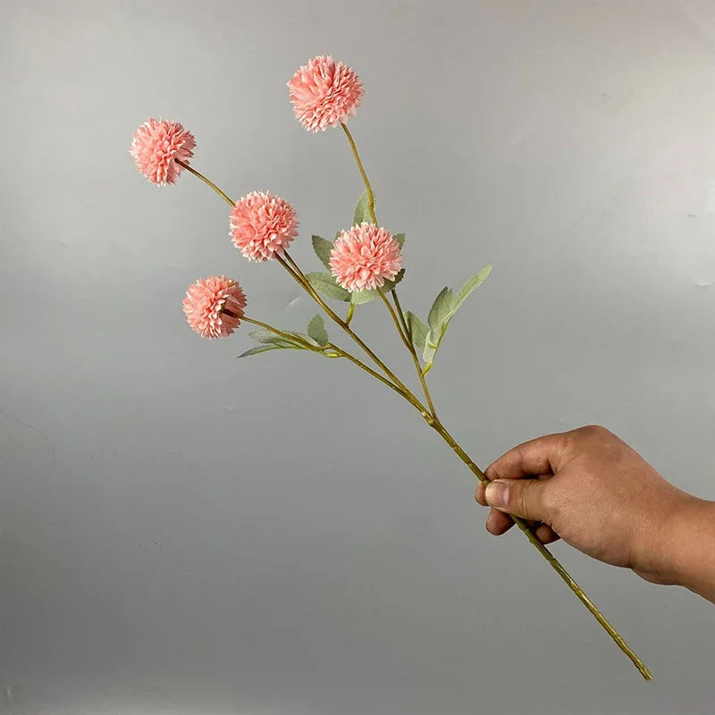 Una mano sostiene un tallo de flores artificiales de crisantemo y diente de león rosadas contra un fondo gris.