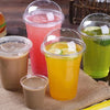 A collection of colorful fruit juices and smoothies in clear disposable plastic cups with lids on a burlap surface.