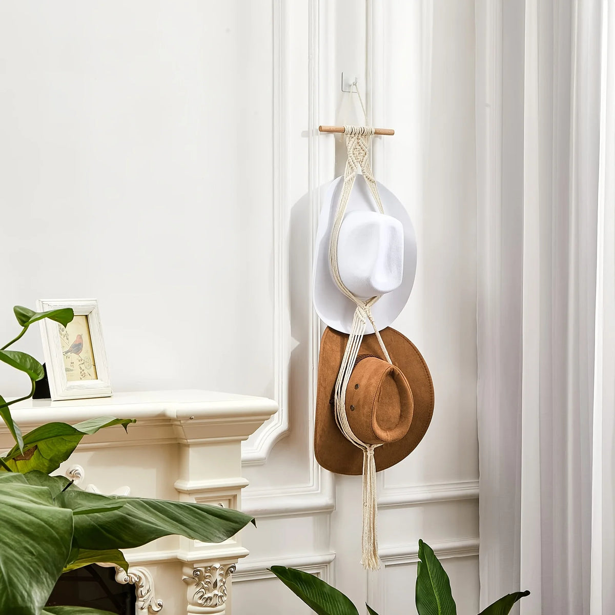 A bohemian macrame hat hanger made of natural fibers is displayed on a white wall, hanging next to a wooden rod.